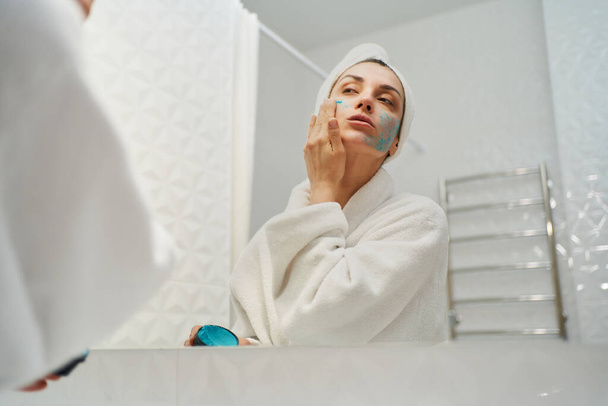 Rejuvenating power of natural skincare. woman wearing a robe while applying a nutritious healthy facial mask and looking in mirror in bathroom - Фото, зображення