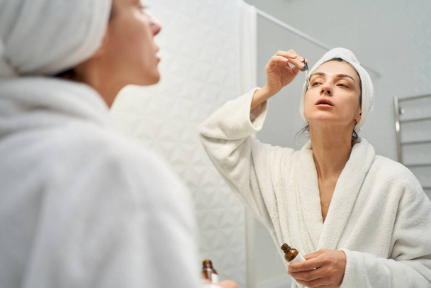 Stunning woman in bathrobe applying serum to her face as part of her daily skincare routine. Perfect for beauty and skincare related projects - Foto, afbeelding