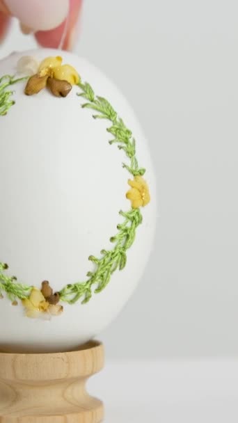 イースターの春の休日料理krashenokはガチョウの卵の女性の手で卵のシェル刺繍を飾る木製のスタンドに完成品を置きます白いマニキュアの光の背景イースターのための手作りの卵 - 映像、動画