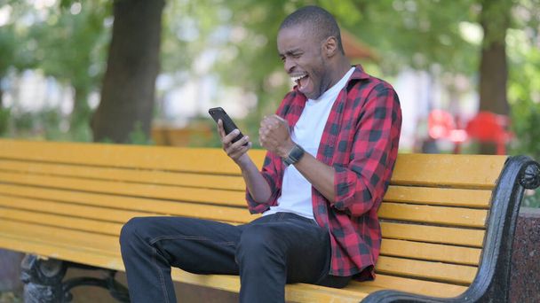 Молодой африканский человек празднует онлайн на смартфоне, сидя на скамейке - Фото, изображение
