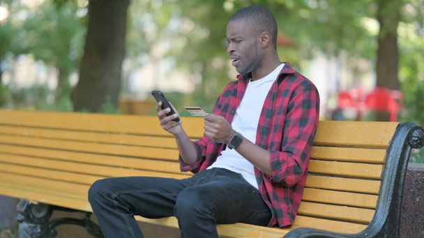 Молодой африканский человек расстроен отказом в оплате онлайн по телефону, сидя на скамейке - Фото, изображение