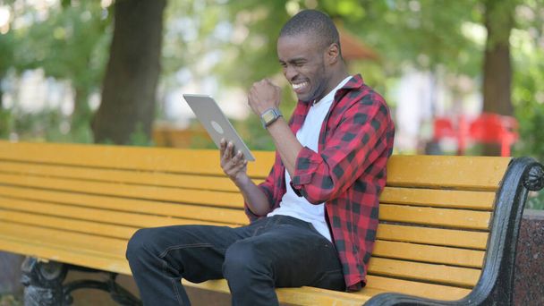 Giovane uomo africano che celebra il successo su tablet mentre si siede all'aperto su una panchina - Foto, immagini