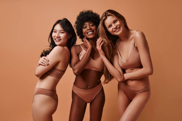 Multi-ethnic multiracial young women in beige lingerie isolated on a beige background. African-American, Caucasian and Asian women posing in the studio. Concept of face body skin care, cosmetology. - Photo, image