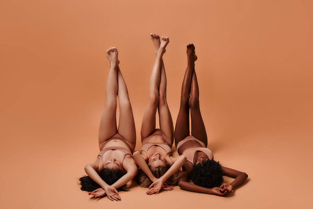 Portrait of beautiful young women of different ethnicities with perfect firm and slim bodies with hairless soft and silky legs crossed isolated on a beige background. - Φωτογραφία, εικόνα