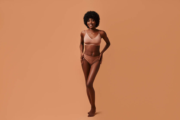 Ritratto di una giovane donna afro-americana in lingerie beige in posa su sfondo beige. Il concetto di cura della pelle, cosmetici e trattamenti termali. - Foto, immagini