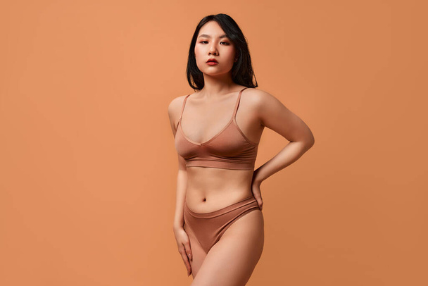 Ritratto di una giovane asiatica in lingerie beige in posa su uno sfondo beige. Il concetto di cura della pelle, cosmetici e trattamenti termali. - Foto, immagini