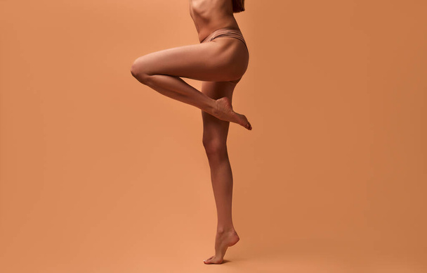 Gesneden portret van de benen van de blanke slanke vrouw in beige lingerie geïsoleerd op een beige achtergrond. Concept van huidverzorging, cosmetologie en spa-behandelingen. - Foto, afbeelding