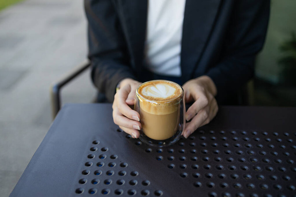 Close-up van jonge vrouw in het bedrijfsleven casual dragen drinken kopje koffie tijdens het zitten aan kleine tafel en genieten van de tijd in de straat cafe. Hoge kwaliteit foto - Foto, afbeelding