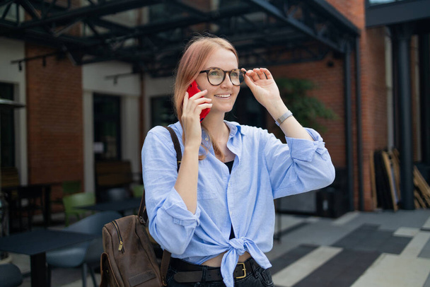 Веселая счастливая стильная женщина с помощью смартфона онлайн приложение, стоя рядом с торговым центром. Счастливая женщина в голубой рубашке идет по улице, глядя на свой смартфон. - Фото, изображение