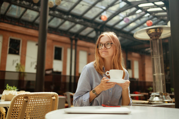 Πορτρέτο του χαμογελαστού Καυκάσου νεαρή γυναίκα σε casual φορούν πόσιμο φλιτζάνι καφέ και χρησιμοποιώντας το κινητό τηλέφωνο, ενώ κάθεται σε μικρό στρογγυλό τραπέζι και να απολαύσετε το χρόνο στο δρόμο καφέ. Υψηλής ποιότητας φωτογραφία - Φωτογραφία, εικόνα