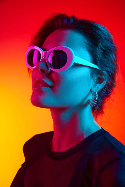 Großaufnahme einer aufgeregten jungen Frau mit Sonnenbrille und schwarzem transparenten Oberteil über gelb-rotem Neonhintergrund. Konzept der Schönheit, Fashion Week, Emotionen, Trends, Werbung - Foto, Bild