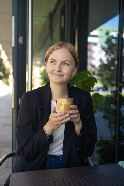 Mosolygó fiatal nő az üzleti alkalmi viselni ivás csésze kávét, miközben ül a kis asztalnál, és élvezi az időt az utcai kávézóban. Kiváló minőségű fénykép - Fotó, kép