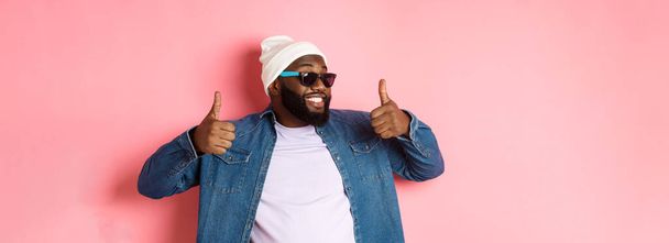 Happy and satisfied african-american guy in sunglasses showing thumbs-up, praise and like promo, agree with you, standing over pink background. - Photo, Image