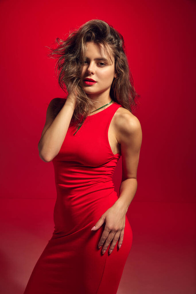 Portret van charmant modemodel, jonge vrouw in trendy kleding, rode strakke midi jurk over bordeaux achtergrond in neon licht. Schoonheidsbegrip, modeweek, menselijke emoties, reclame - Foto, afbeelding