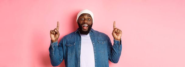 Cheerful Black man showing fantastic promo offer, pointing fingers up and smiling amused, standing over pink background. - Photo, Image