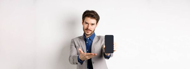 E-commerce en online shopping concept. Man demonstreerde lege smartphone scherm voor u, tonen telefoon display om iets te introduceren, staan op een witte achtergrond. - Foto, afbeelding
