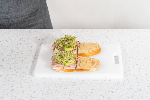 Assembling ham, cucumber, and sprout sandwiches on the white cutting board. - Zdjęcie, obraz