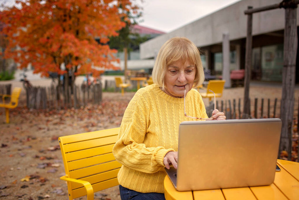 Femme active à la fin de la soixantaine, assise sur un banc de parc, entourée de couleurs automnales. Elle utilise habilement son ordinateur portable. Adaptabilité à l'ère numérique, une technologie de pointe et moderne - Photo, image