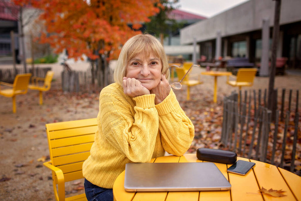 60年代後半の思慮深い女性は,秋の色に囲まれた公園のベンチに座っています. 彼女はラップトップを巧みに使っている. デジタル時代の適応可能性,技術に精通したシニアと現代技術 - 写真・画像