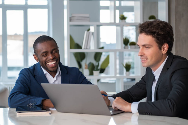 Cheerful African American businessman wearing formal suit talking to colleague discussing working moments at office workplace with laptop in background. Concept of teamwork, cooperation, coworking - Foto, Imagem