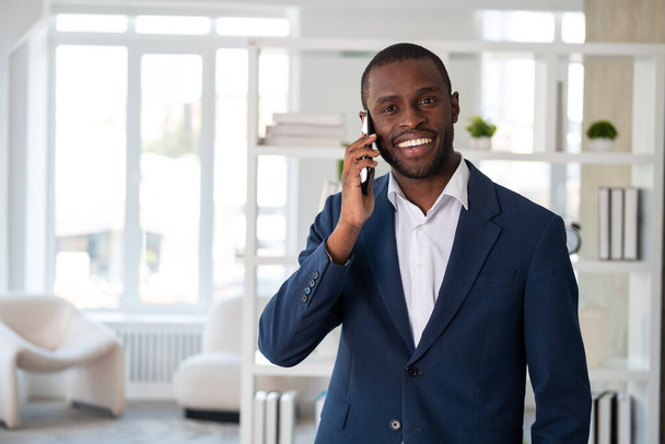 Smiling African American businessman wearing formal suit is standing talking on smartphone looking at camera at office workplace. Concept of model, business person, time management, device, gadget - Foto, Imagem