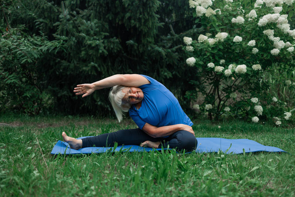 Sportieve vijftiger jaren senior vrouw poseert op yoga mat buiten gekleed in sport kleding alleen in het park of bos in de natuur. Hoge kwaliteit foto - Foto, afbeelding