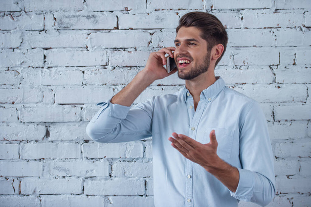 Handsome young businessman in classic shirt is talking on the mobile phone and smiling, standing in front of white brick wall - Photo, Image