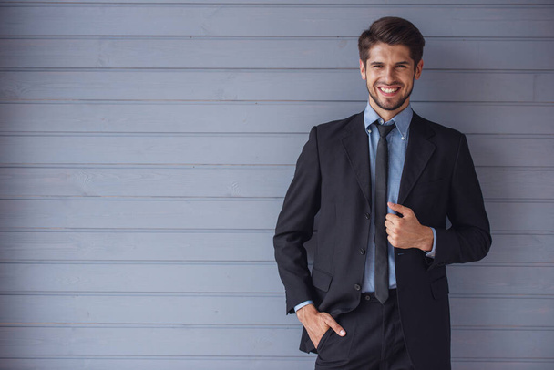 Guapo joven empresario confiado en traje clásico está mirando a la cámara y sonriendo, de pie frente a la pared gris
 - Foto, imagen