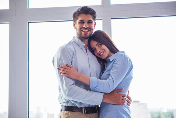Beautiful young business couple is hugging, looking at camera and smiling, standing before window - Photo, Image