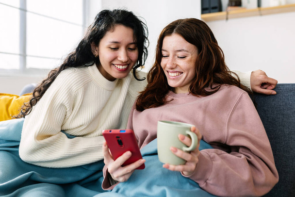 Happy young female friends using mobile phone sitting together on sofa at home. Winter domestic lifestyle and roommate people concept - Fotó, kép