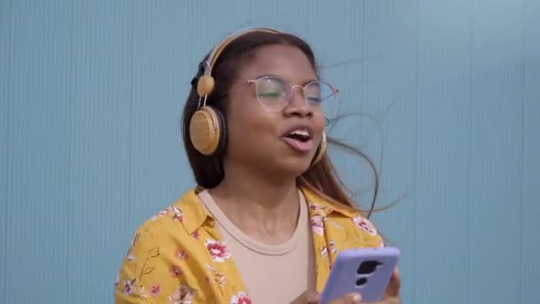 Vidám fiatal afro-amerikai nő élvezi a mobil zene tánc és éneklés. Boldog mosolygós lány mobiltelefonnal és fejhallgatóval. Generation z szabadidős női emberek kék hátterű.  - Felvétel, videó