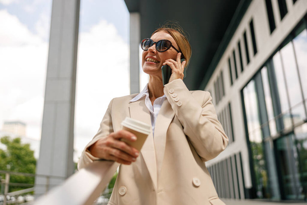 Улыбающаяся женщина-офисный работник стоит на современном фоне здания и пьет кофе - Фото, изображение