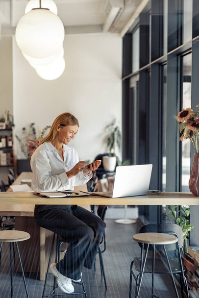 Женщина менеджер по продажам держит телефон, сидя в современном коворкинге и работая на ноутбуке - Фото, изображение