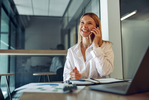 居心地の良いオフィスに座って電話で話している間,ラップトップで働く笑顔の成熟したビジネス女性 - 写真・画像