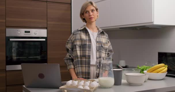 smiling happy young woman is preparing breakfast for husband in the morning at home Slow motion blonde girl in casual clothes looking at camera, standing behind the table - Video, Çekim