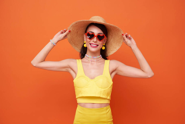 Joyeux attrayant femme en robe d'été et chapeau posant tout en se tenant debout et en regardant la caméra isolée sur fond orange. - Photo, image