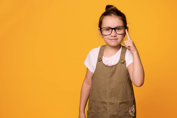 Suloinen pikkutyttö, ensiluokkainen, alakoululainen, jolla on opettajan silmälasit, hymyilee kameran edessä, osoittaa sormellaan, eristettynä oranssin taustan päälle kopioimalla mainostilaa.. - Valokuva, kuva