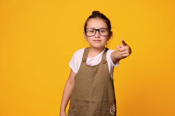 Serious primary school student girl, first grader wearing teacher's black framed glasses, points her index finger at camera, showing warning hand sign, isolated orange background. Back to school - Photo, Image