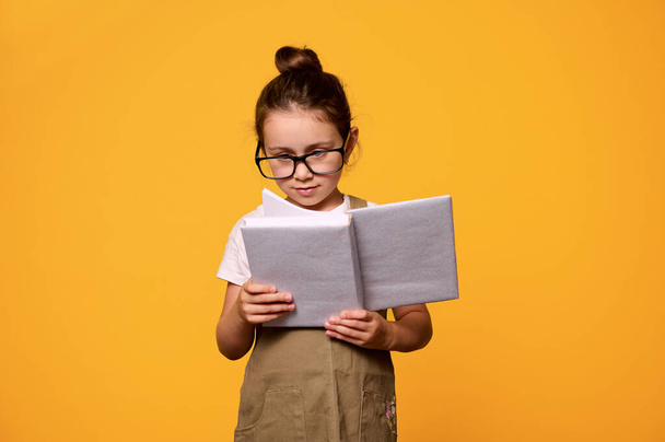 Kluges Grundschülermädchen mit Brille, liest ein Buch, orangefarbener Hintergrund. Menschen. Aufruhr. Welttag des Buches. Lehrertag. Zum neuen Semester zurück in die Schule - Foto, Bild