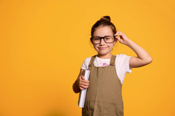 Καυκάσιος μικρό όμορφο κοριτσάκι, μια μαθήτρια της πρώτης τάξης κρατώντας ένα βιβλίο, χαμογελώντας κοιτάζοντας την κάμερα μέσα από τα κομψά γυαλιά της, απομονωμένη πάνω από πορτοκαλί φόντο στούντιο με δωρεάν διαφημιστικό χώρο. - Φωτογραφία, εικόνα