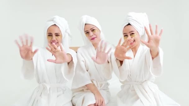 Drei junge asiatische Frauen in Bademänteln, Handtücher auf dem Kopf, unterhalten sich gern und probieren Augenpolstermasken aus. Freundinnen haben Spaß und winken bei Wellness-Behandlungen im Luxushotel. - Filmmaterial, Video