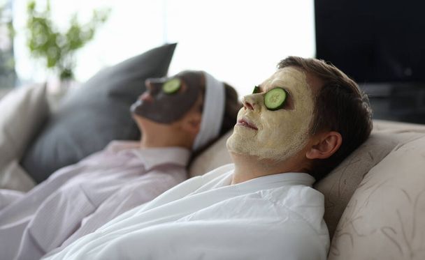 Portrait of couple enjoying beauty treatment on sofa at home. Man and woman applying facial clay masks and moisturizing slices of cucumber. Wellness and skincare concept - Photo, Image