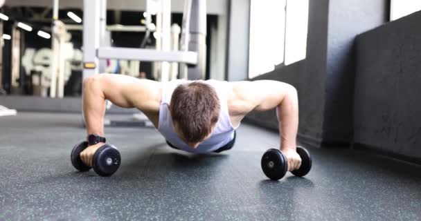 Jeune homme musclé fait push-ups s'appuyant sur des haltères dans les mains. Homme améliore la forme physique de travail dans la salle de gym. Mode de vie sain ralenti - Séquence, vidéo