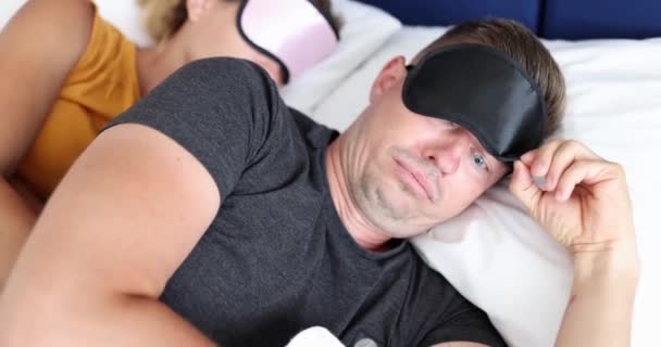 Młody człowiek zdejmuje czarną maskę snu leżącą rano na łóżku. mężczyzna otwiera usta z zaskoczony wyraz twarzy patrząc na kobiety leżące w pobliżu - Materiał filmowy, wideo