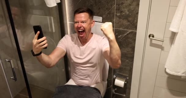 Muž šťastný vyhrát hru v smartphonu sedí na záchodové míse ve světle koupelny bydlení. Koncept vítězného radosti a přirozeného zpomalení procesu - Záběry, video