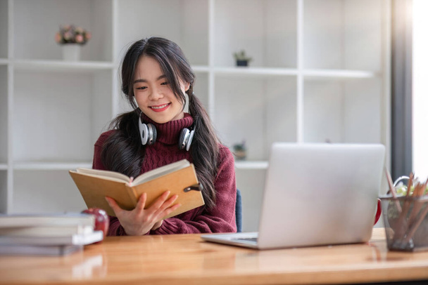 Aziatische vrouwelijke student studeren online met behulp van koptelefoon en laptop het maken van notities in notebook zitten aan huis tafel ontspannen en het veranderen van plaatsen om school te ontspannen.. - Foto, afbeelding