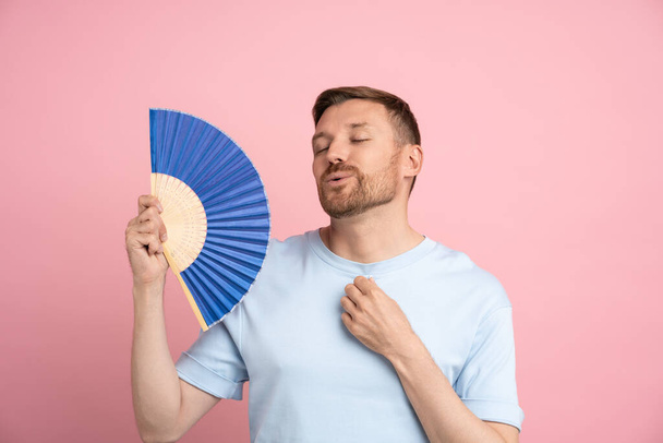 Πορτρέτο παιχνιδιάρικο κουρασμένος άνθρωπος κυματίζει μπλε ανεμιστήρα πάσχει από stuffiness στέκεται σε ροζ φόντο. Υπερθέρμανση υψηλής θερμοκρασίας, ζεστό καλοκαίρι έννοια του καιρού. Μπάνερ, αφίσα ποζαρίσματος καλλιτεχνικού αρσενικού. - Φωτογραφία, εικόνα