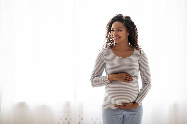 Χαμογελώντας μαύρη έγκυο γυναίκα αγκαλιάζει μεγάλη κοιλιά και κοιτάζοντας πέρα από το χώρο αντιγράφων, όμορφη νεαρή Αφροαμερικανή προσδοκώμενη γυναίκα στέκεται κοντά στο παράθυρο στο σπίτι, απολαμβάνοντας το χρόνο εγκυμοσύνης - Φωτογραφία, εικόνα