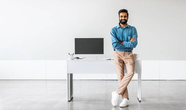 Fiducioso uomo d'affari indiano seduto sulla scrivania in posa con le braccia incrociate che mostra computer PC con schermo vuoto in ufficio, mockup, modello. Persone, tecnologia, concetto di lavoro a distanza - Foto, immagini