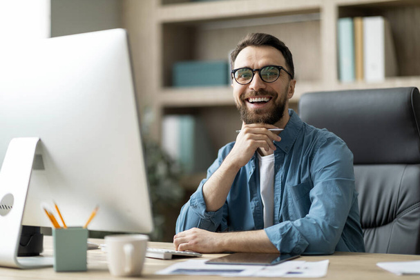 Modern Ceo. Portrait Of Handsome Young Businessman Sitting At Desk In Office, Happy Young Male Entrepreneur In Eyeglasses Posing At Workplace, Looking And Smiling At Camera, Free Space - Photo, Image
